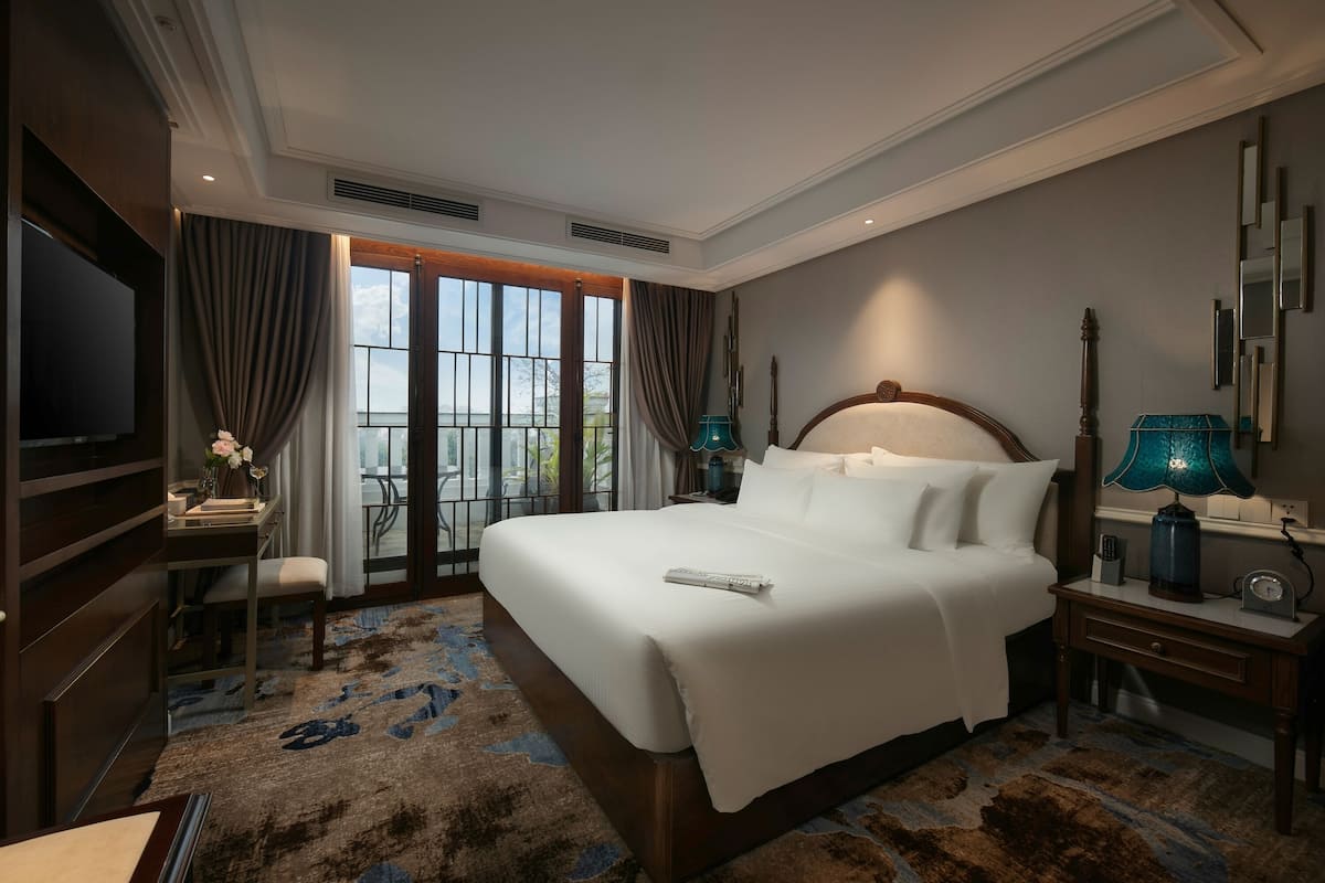 Habitación de hotel con una cama grande, un escritorio con silla, televisión y vistas al exterior a través de amplias ventanas.