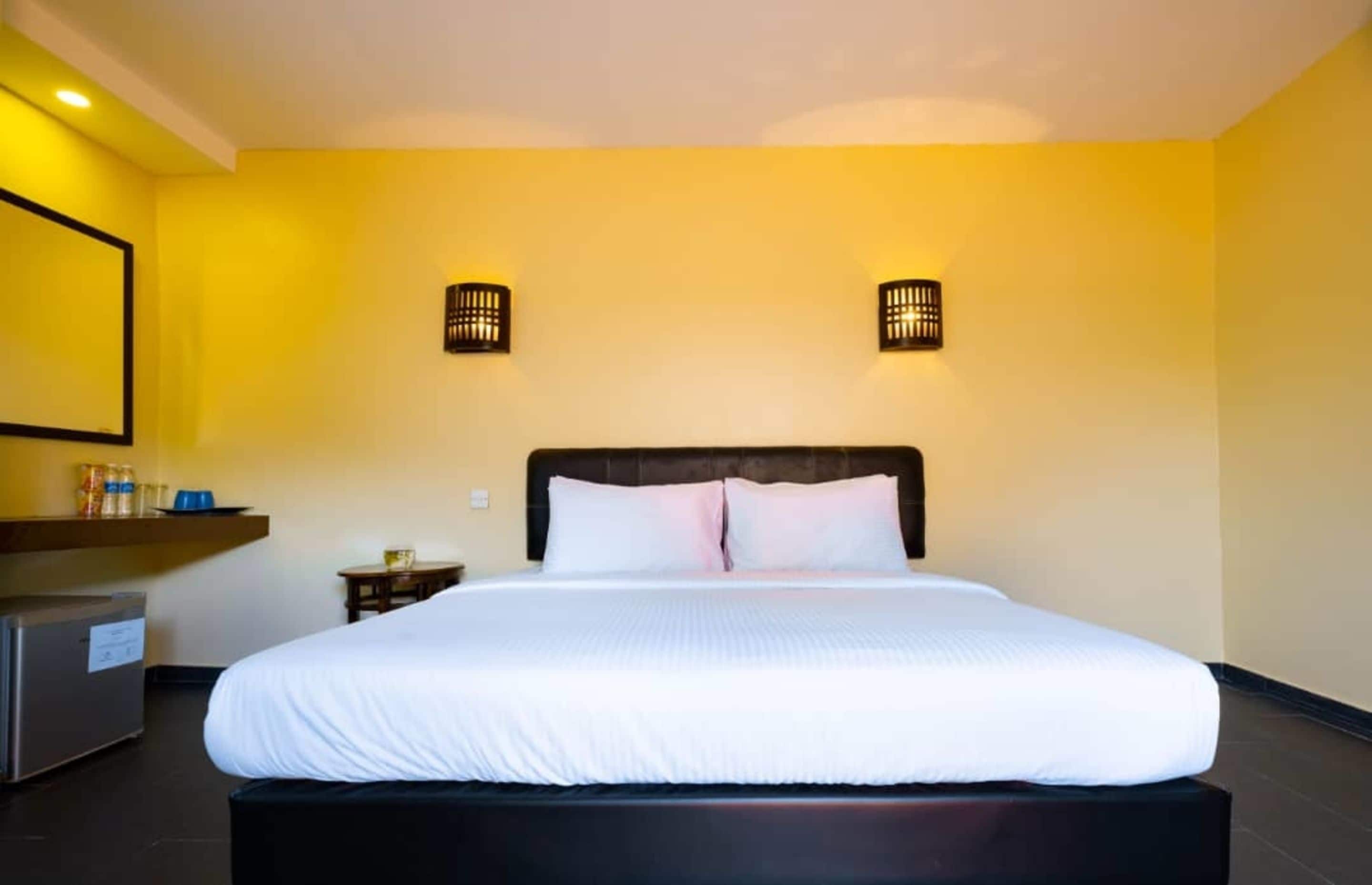 Une chambre d’hôtel avec un grand lit, une petite table de chevet, une commode et deux lampes fixées au mur.
