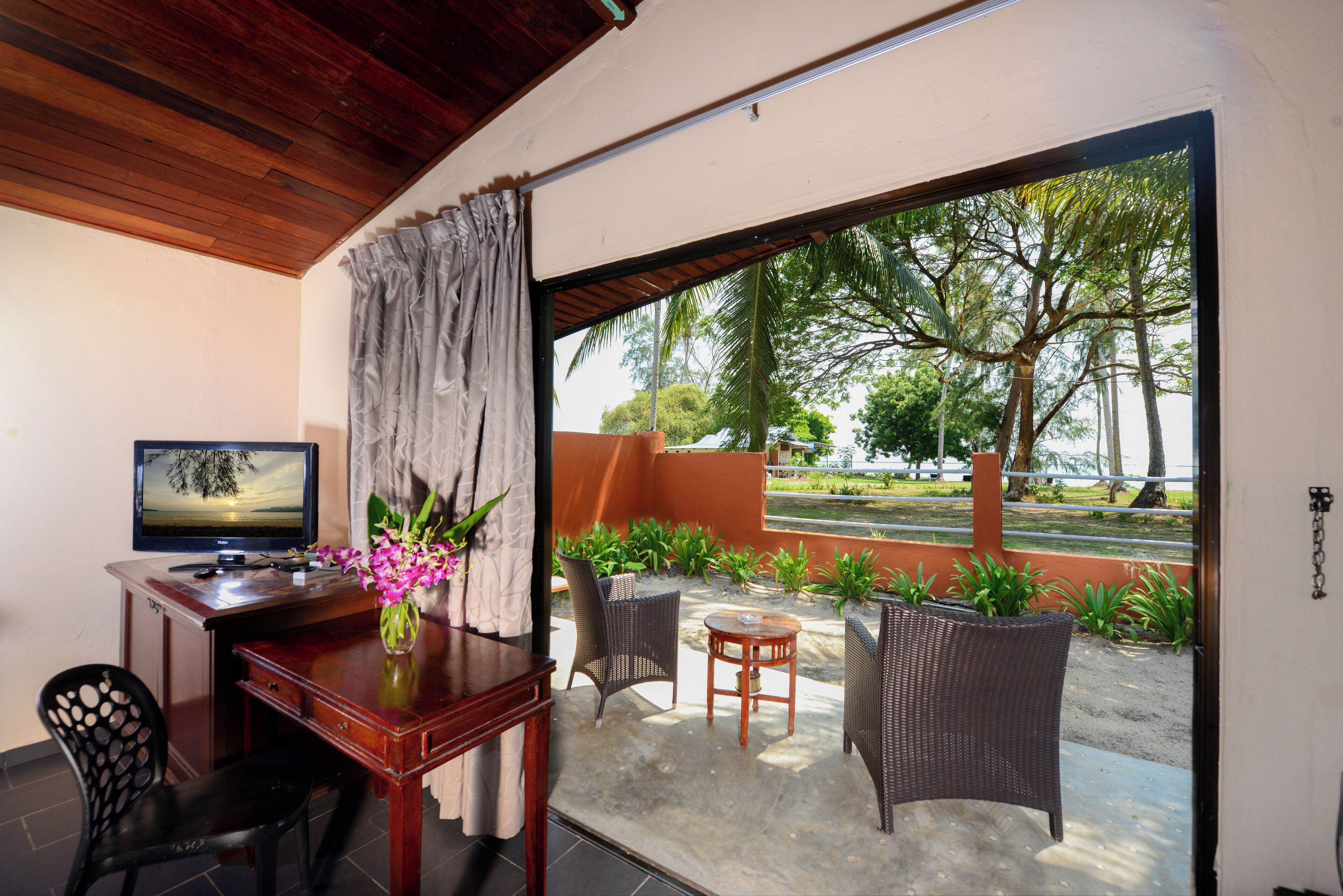 A room with a wooden desk, a television, wicker chairs, and a view of a landscaped garden.