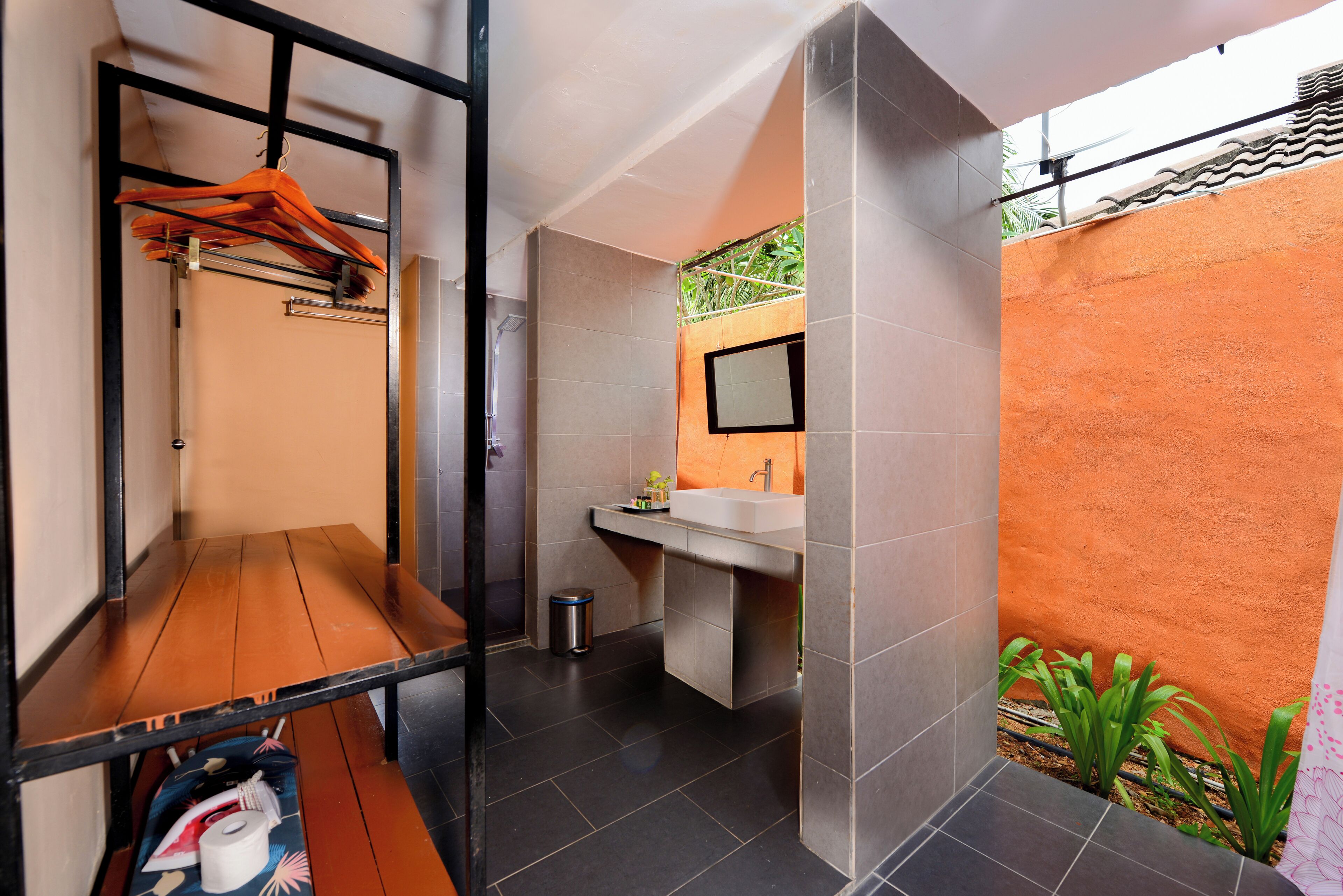 A compact bathroom with a wooden bench, a hanging clothes rack, a sink, and a trash bin.