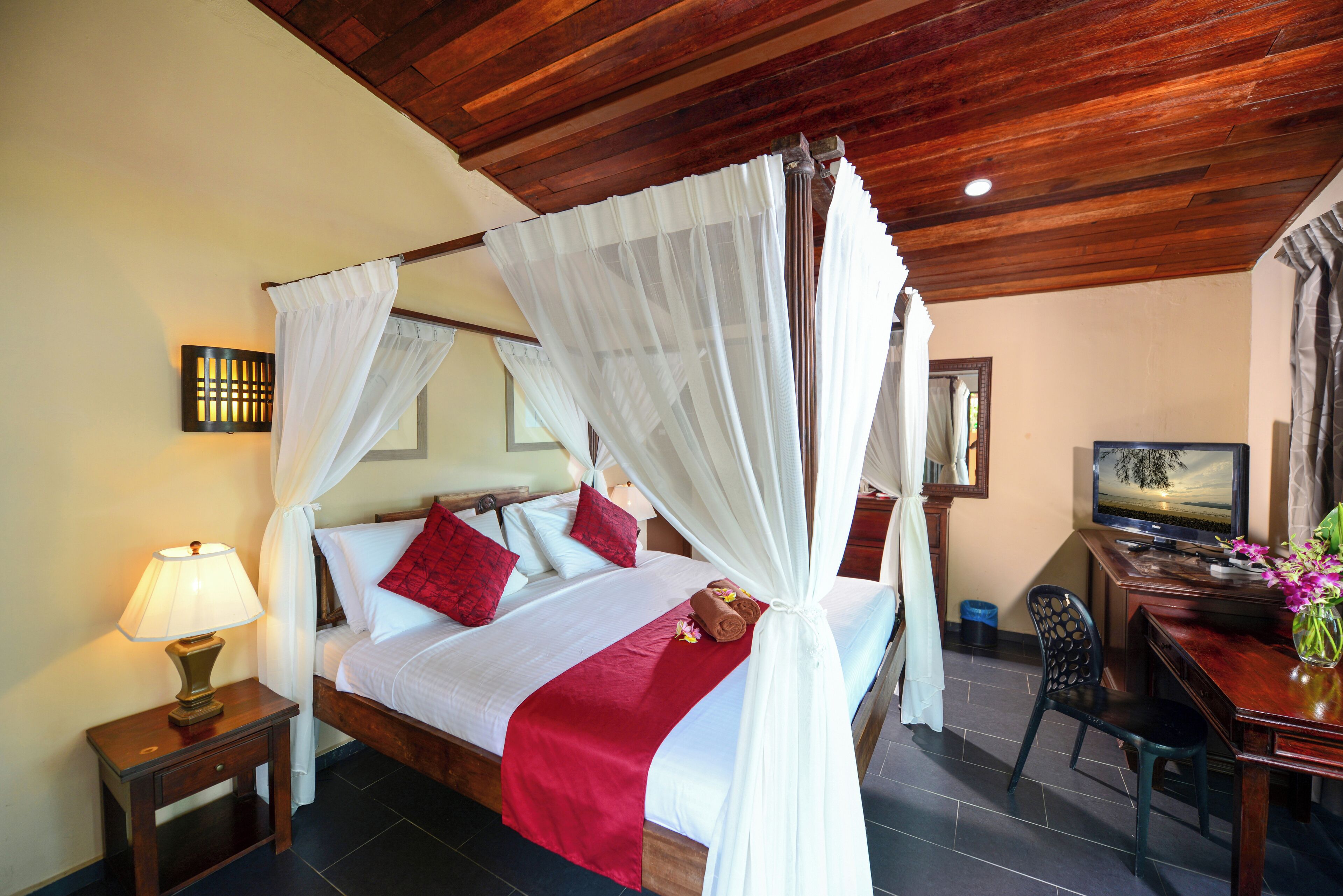 A four-poster bed with white and red bedding, a bedside table with a lamp, and a desk with a computer and vase of flowers.