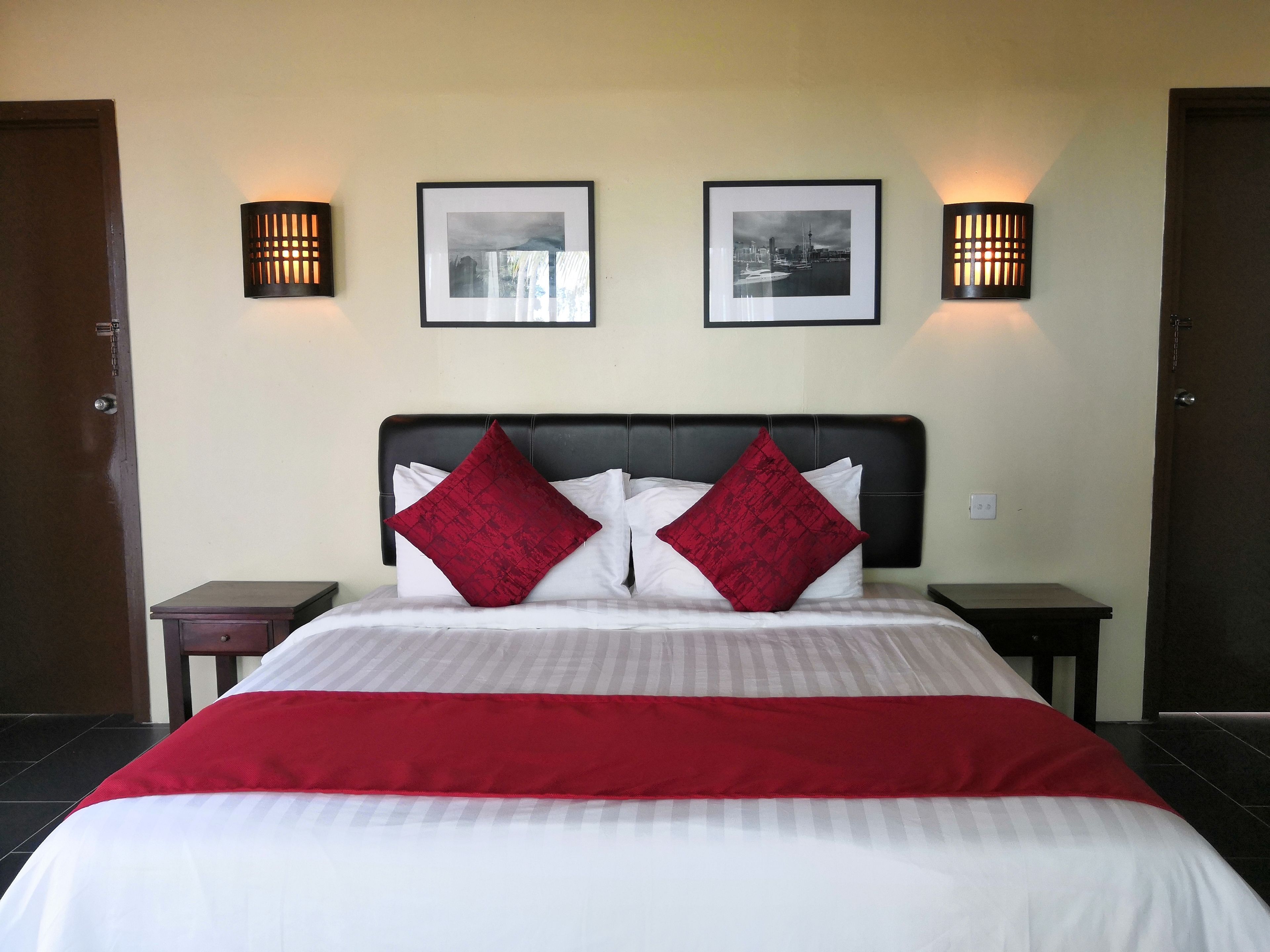 A neatly made bed with a red and white bedspread, two red pillows, and a black headboard. There are two framed pictures on the wall, and two wall-mounted lamps on either side of the bed.