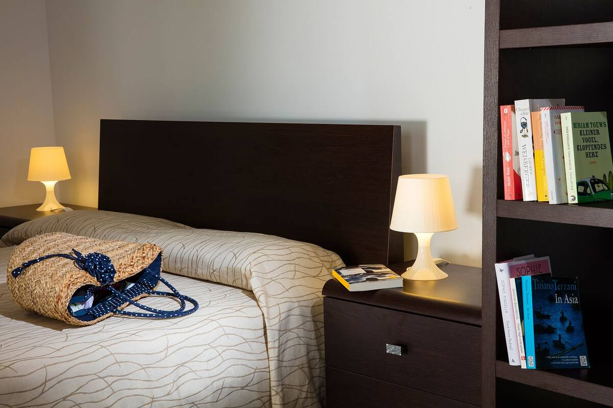 A neatly made bed with a woven bag and a book, a bedside lamp, and a bookshelf with several books.