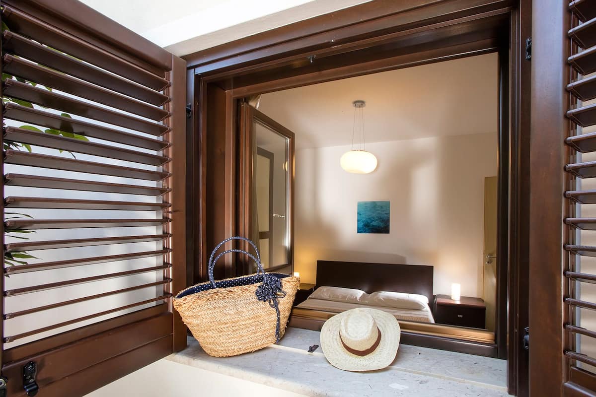 A room with a bed, a nightstand, a lamp, a woven basket, and a straw hat on the windowsill.