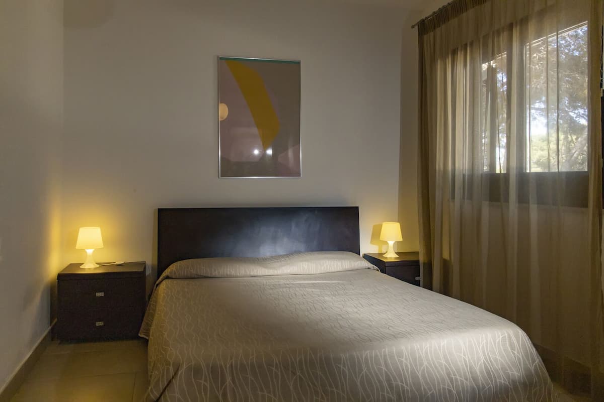 A hotel room with a large bed, two bedside tables with lamps, a wall-mounted artwork, and a window with curtains.