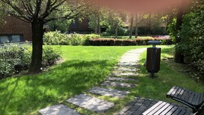 Een geplaveid pad dat leidt naar een zitgedeelte met een vuilnisbak, omringd door groen en een gebouw op de achtergrond.