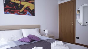 Een net opgemaakt bed met witte en paarse kussens, een bedlampje en een houten deur.