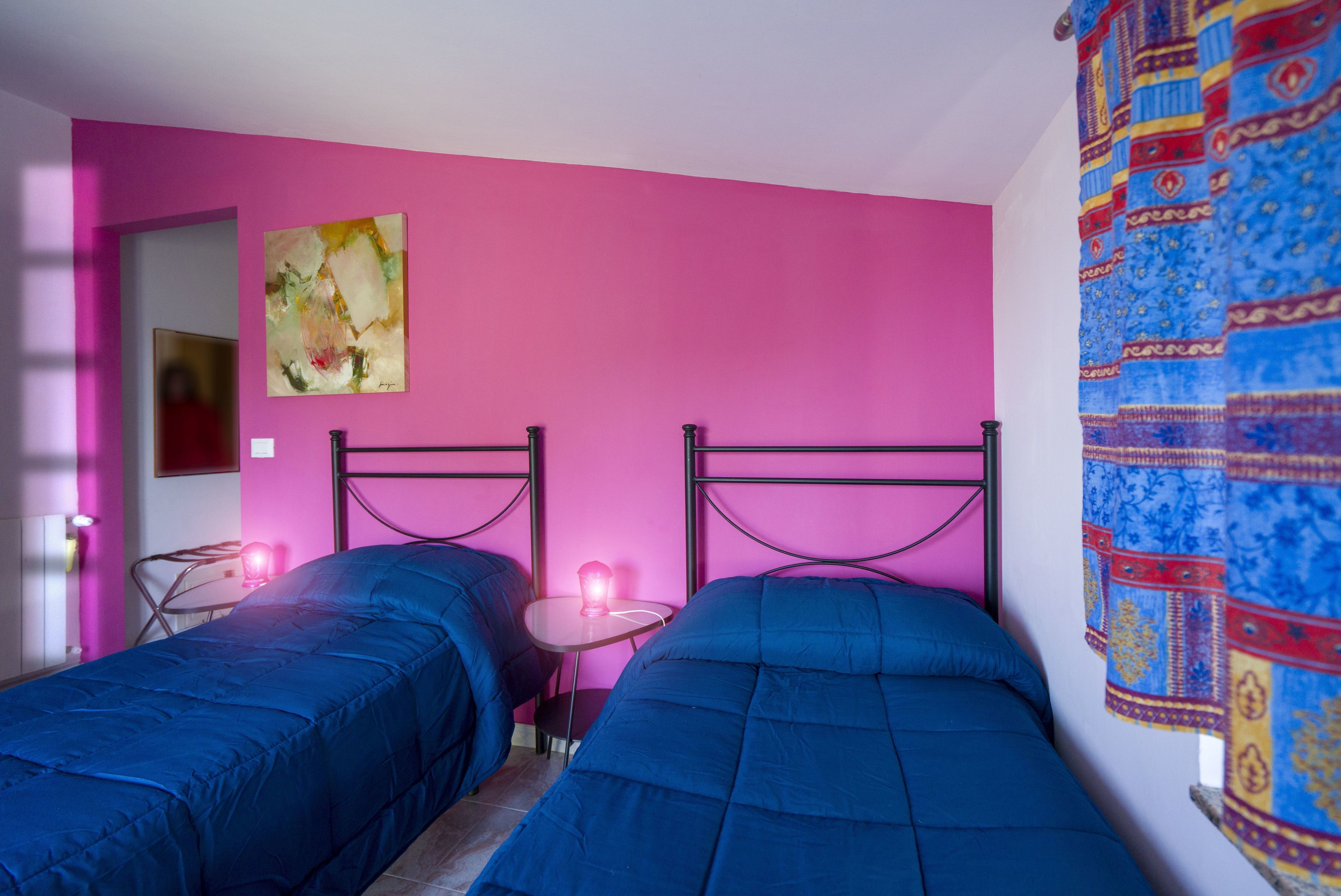 Una habitación con dos camas, una pared rosa, una mesita con una lámpara rosa y una cortina colorida.