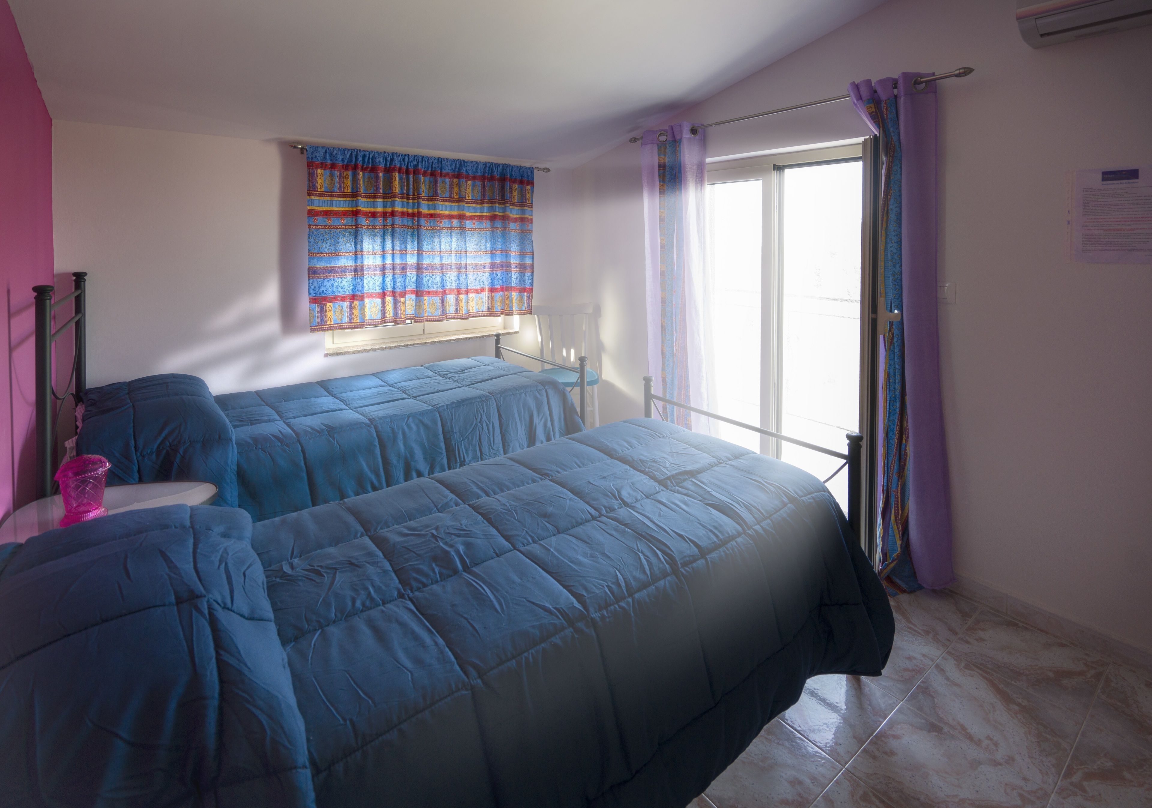A bedroom with two beds, a window with curtains, a bedside table, and a wall-mounted air conditioner.