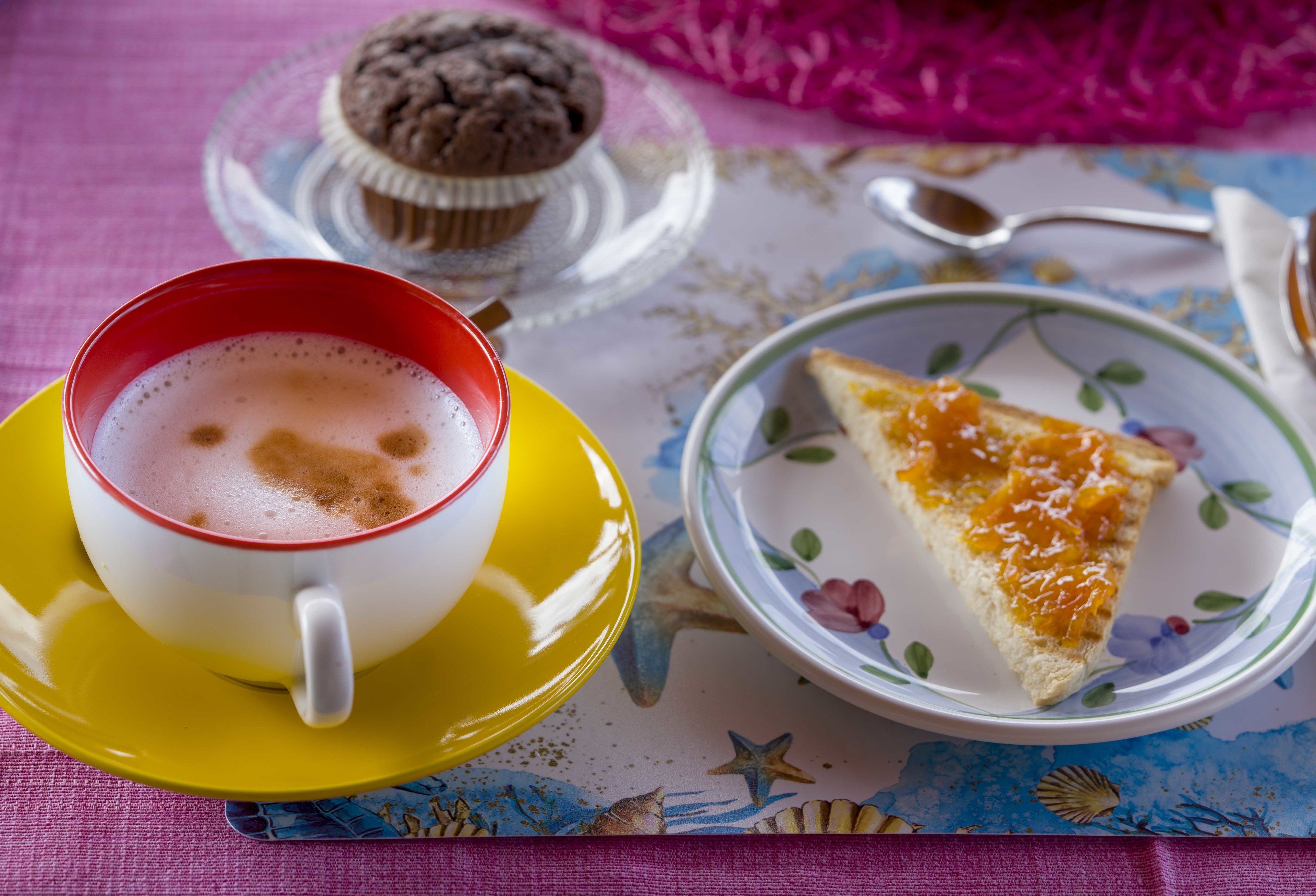A cup of coffee on a yellow saucer, a slice of orange cake on a floral plate, and a muffin on a glass stand.