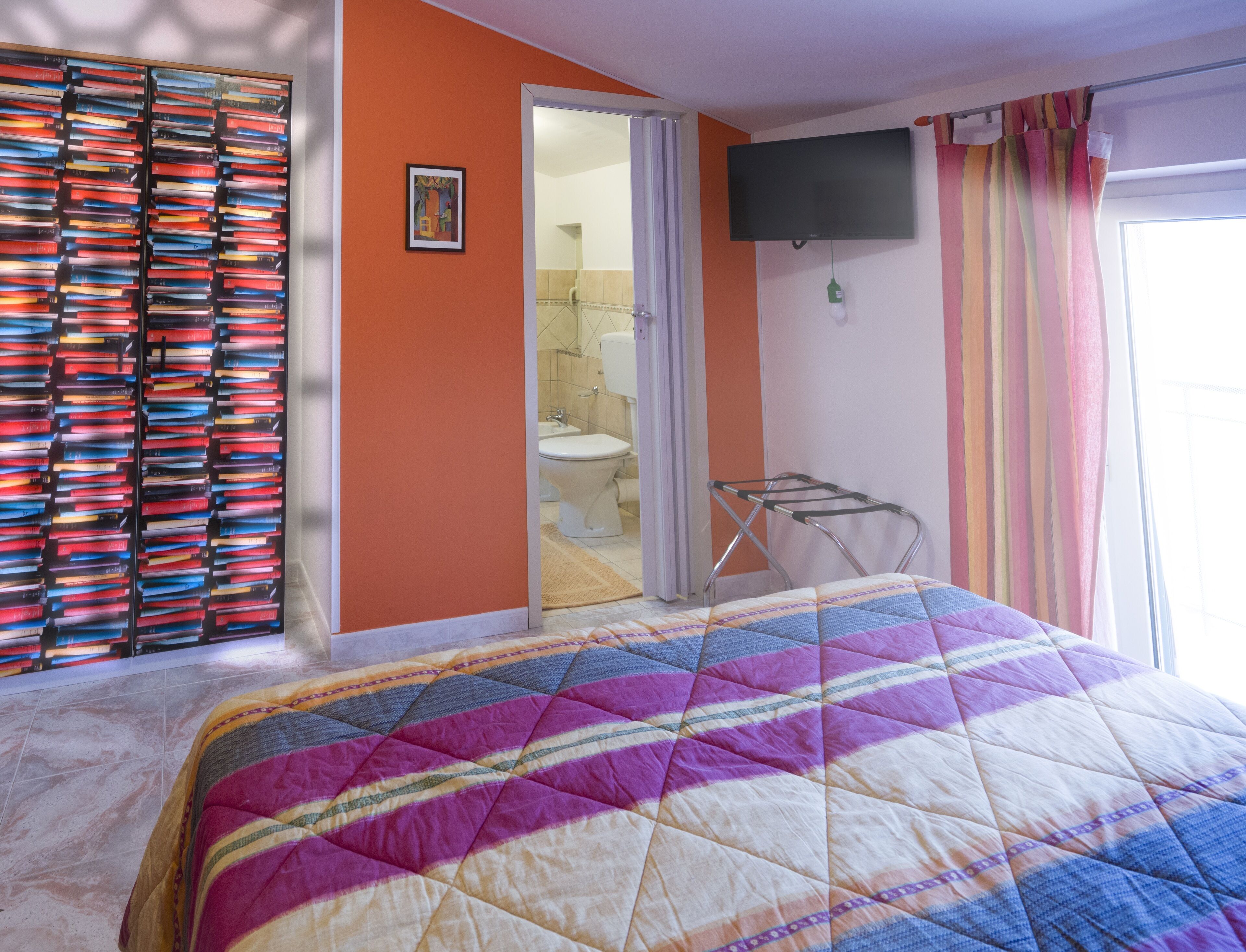 Une chambre à coucher avec un lit, une bibliothèque, un téléviseur et une salle de bain visible à travers une porte ouverte.