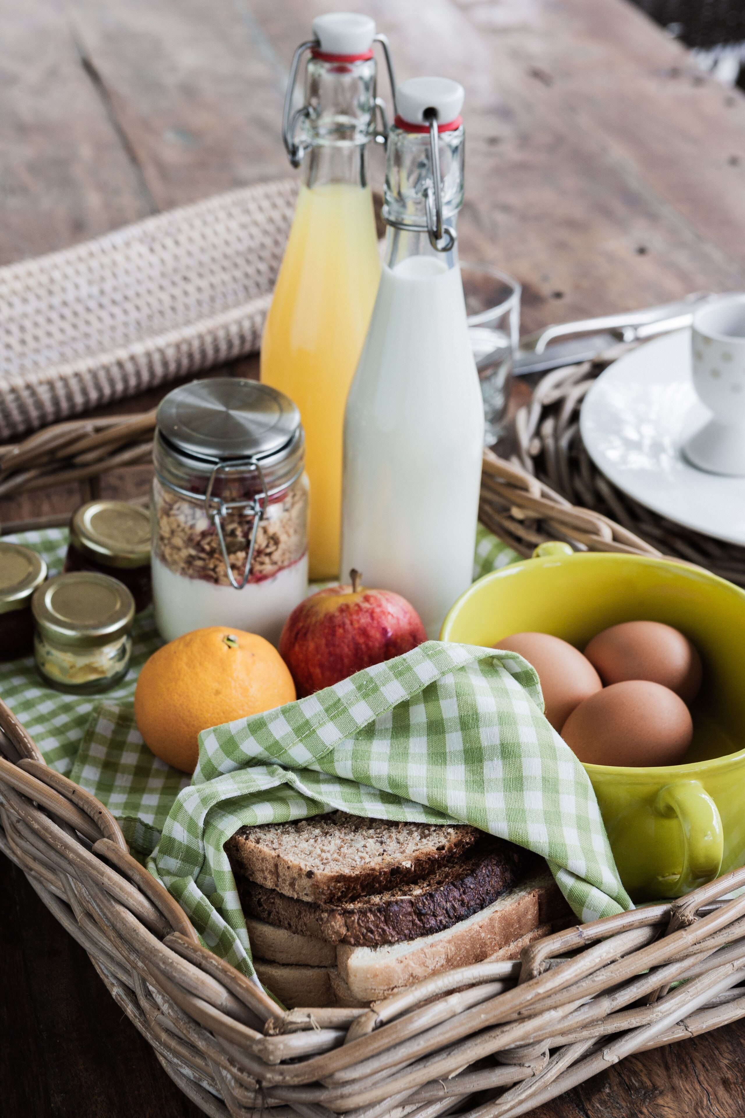 A wicker tray with breakfast items including bread, eggs, an apple, a glass of orange juice, and a bottle of milk.
