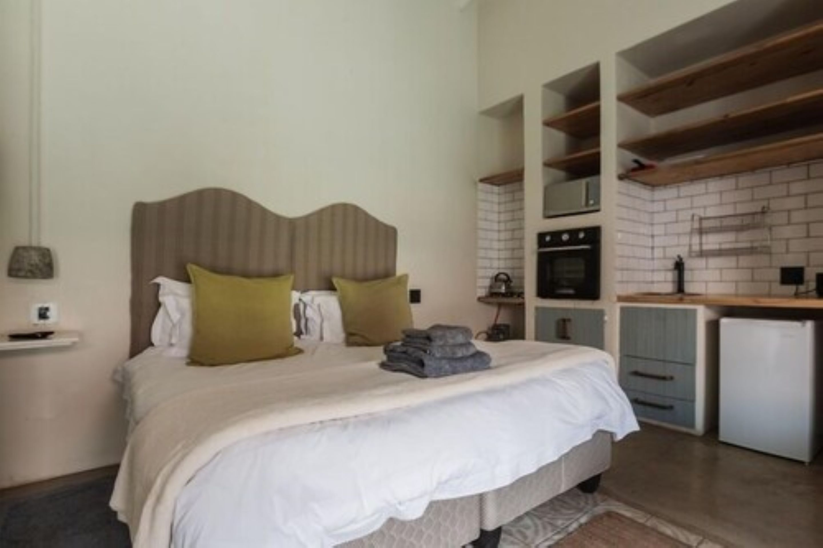 A bedroom with a bed, pillows, a small bedside table, and a nightstand. Adjacent to the bedroom is a kitchenette with a refrigerator, oven, and a countertop.