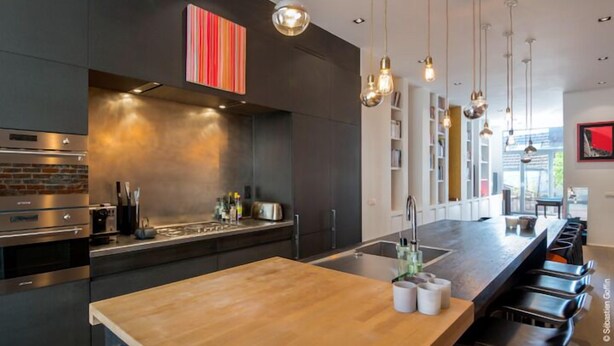 Une cuisine moderne dotée d’un îlot central, d’appareils électroménagers en acier inoxydable et de luminaires suspendus.
