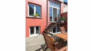 Un bâtiment rouge avec un escalier en bois, un balcon avec une table et des chaises, et un pot de fleurs posé sur le rebord de la fenêtre.