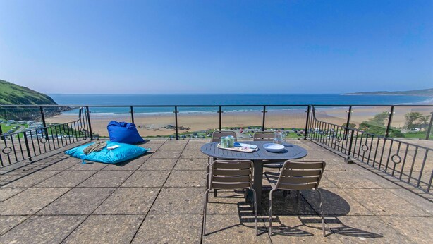Une terrasse avec une table et des chaises, un pouf bleu et une vue sur la plage et l’océan.