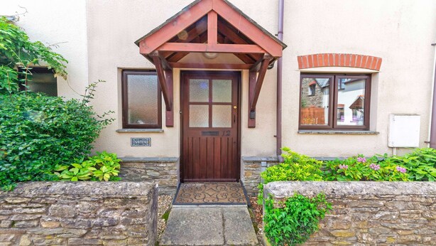 Une maison avec une porte d’entrée en bois, un mur en pierre et un petit jardin avec des plantes.