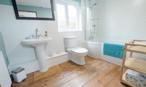Ein Badezimmer mit weißem Waschbecken, Toilette und Badewanne. Über der Toilette befindet sich ein Fenster und es gibt einen Duschbereich mit Glastür. Eine Holzablage mit blauem Handtuch ist an der Wand angebracht.