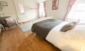 Ein Schlafzimmer mit Holzboden, einem Bett mit weißer und grauer Bettwäsche, einem Sessel mit grauem Kissen, einer weißen Kommode und einem Spiegel.