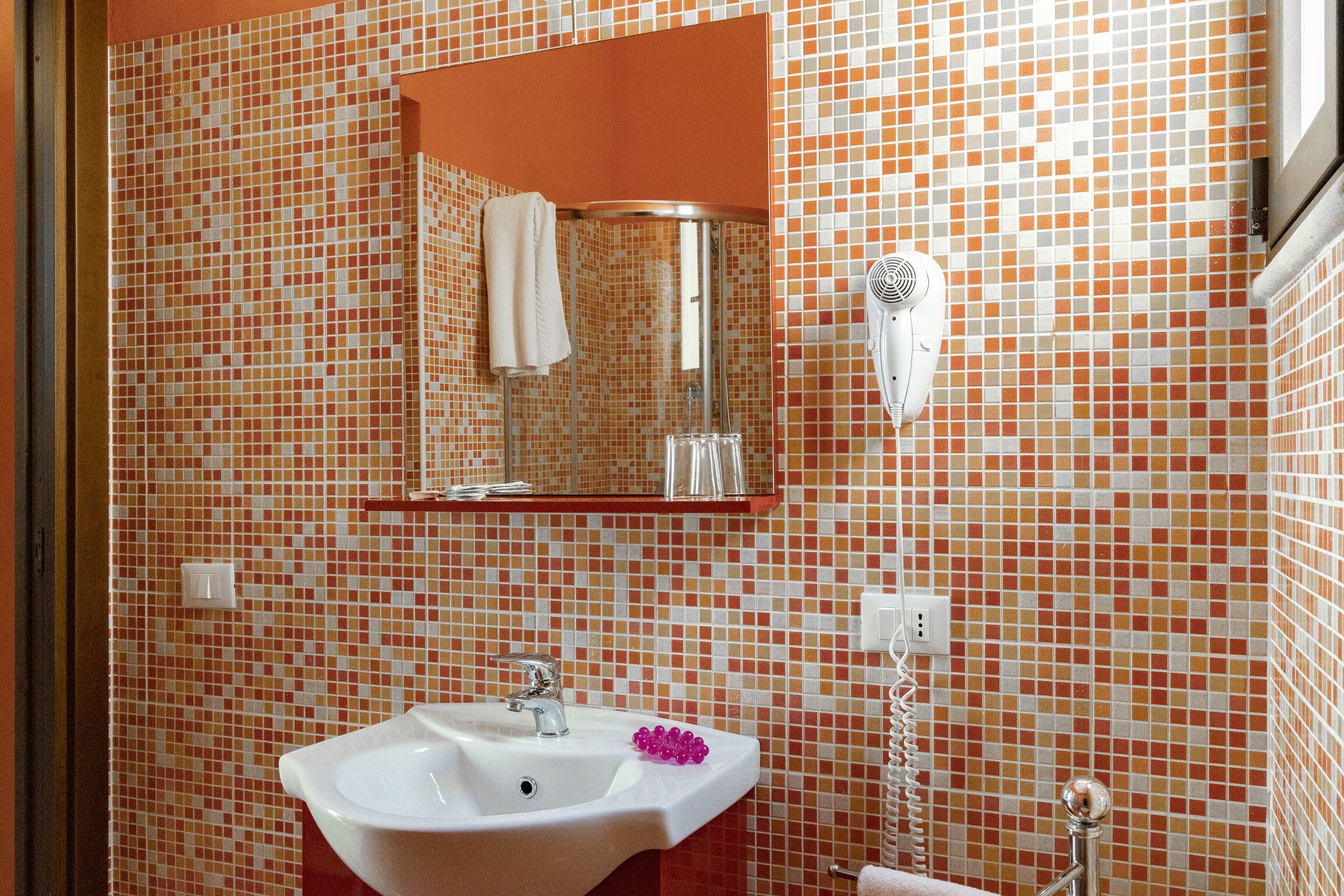 Ein Badezimmer mit einer mosaikverkleideten Wand, einem Waschbecken mit Spiegel darüber und einem an der Wand montierten Föhn.
