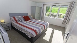 A bedroom with a bed, a nightstand, a wardrobe, and a window with a view of a lawn. - Clifton Court Apt 19 With Heated Pool (Braunton)