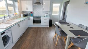 A modern kitchen with white cabinets, a built-in oven, a microwave, and a sink. There is a dining table with chairs and a window providing natural light.