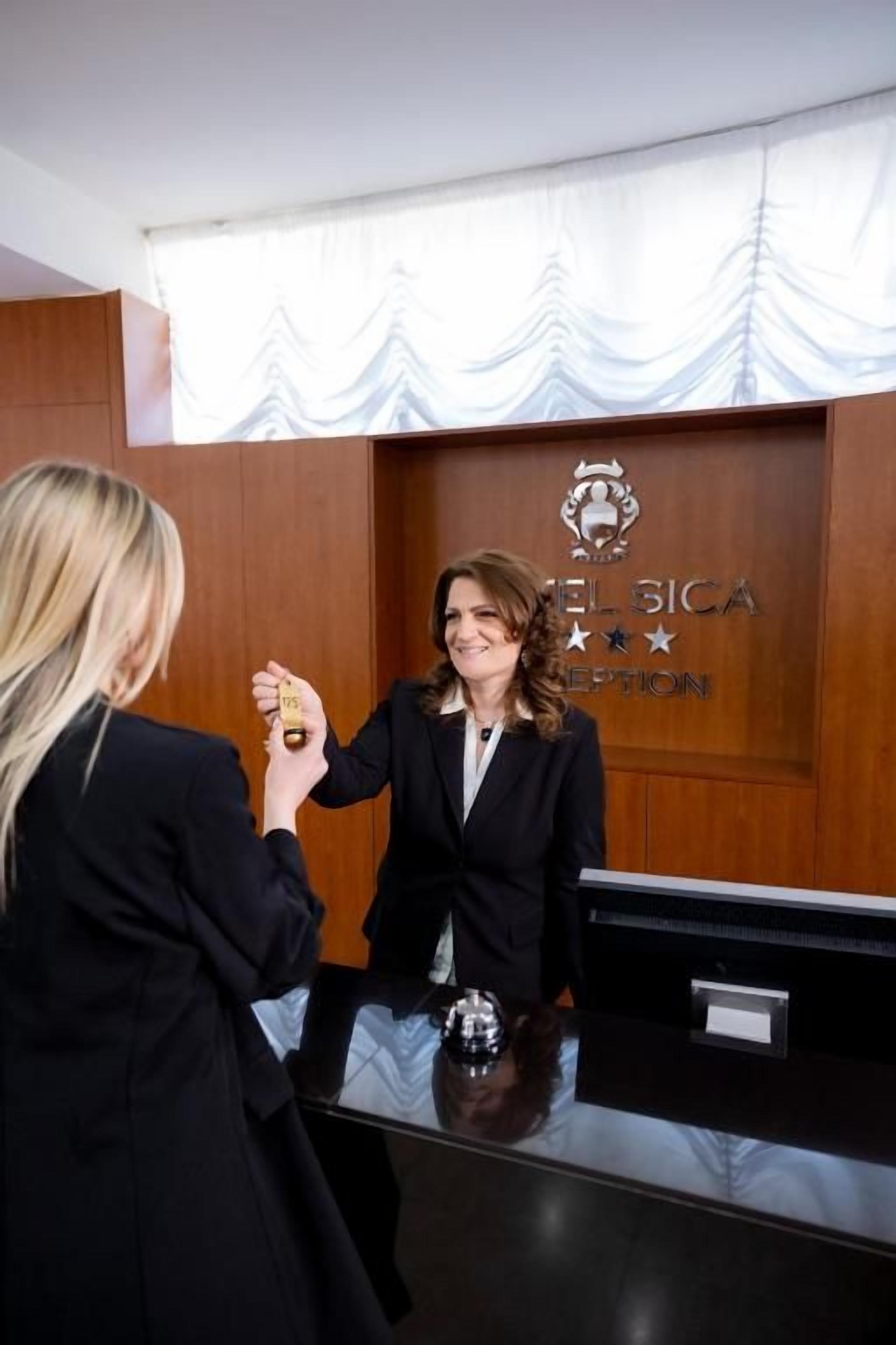 A reception desk with a wooden panel and a logo, a person handing over a key, and a glass counter.