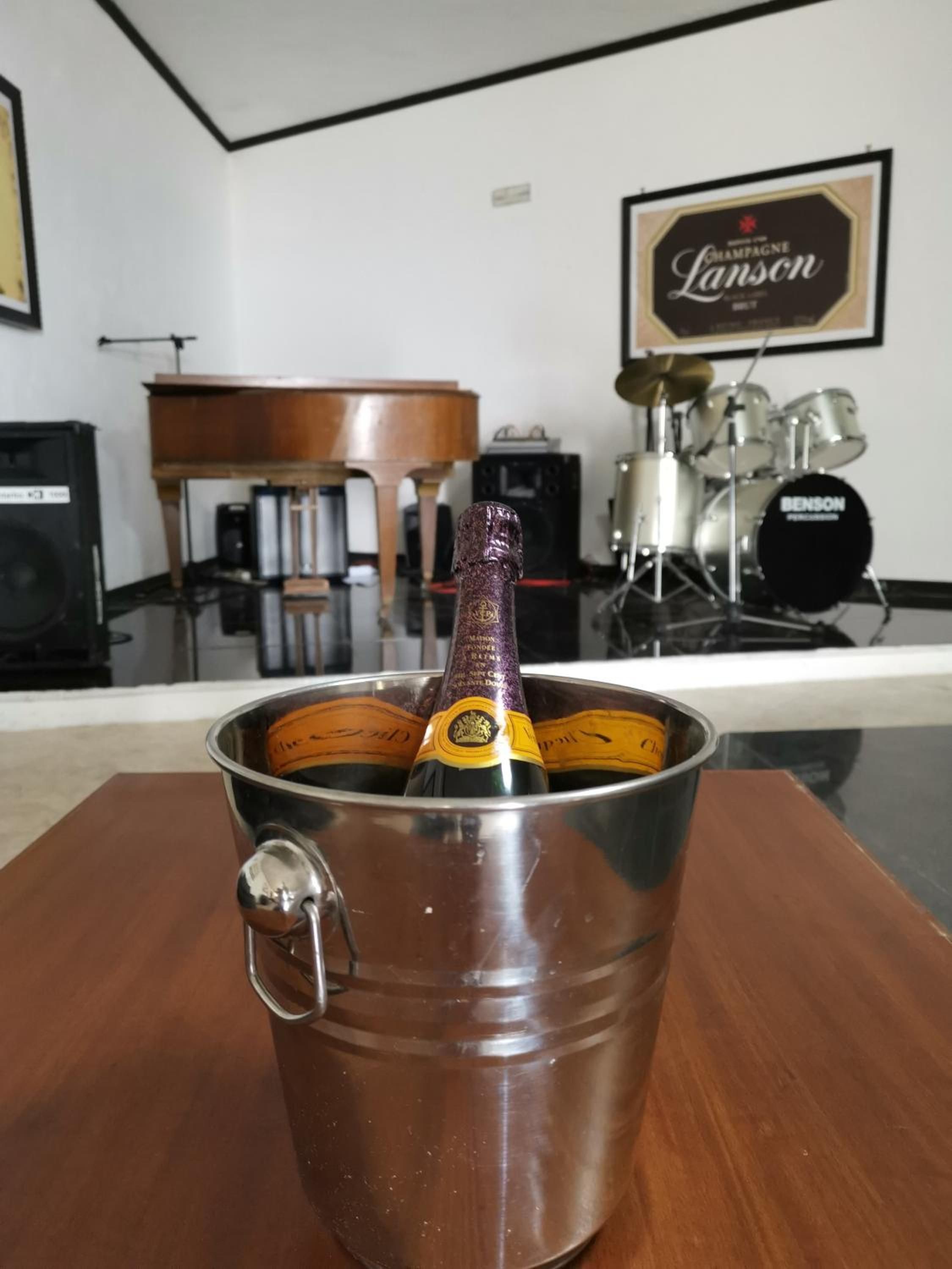 A metal bucket with a bottle of champagne, a wooden table, a piano, and a drum set in the background.
