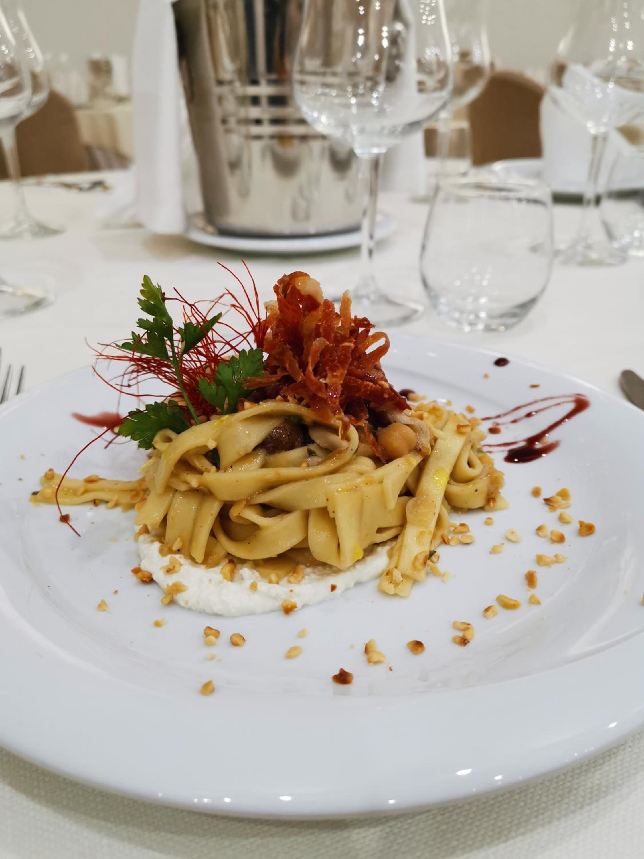 A plated pasta dish with garnishes, accompanied by a white plate and a silver pitcher in the background.