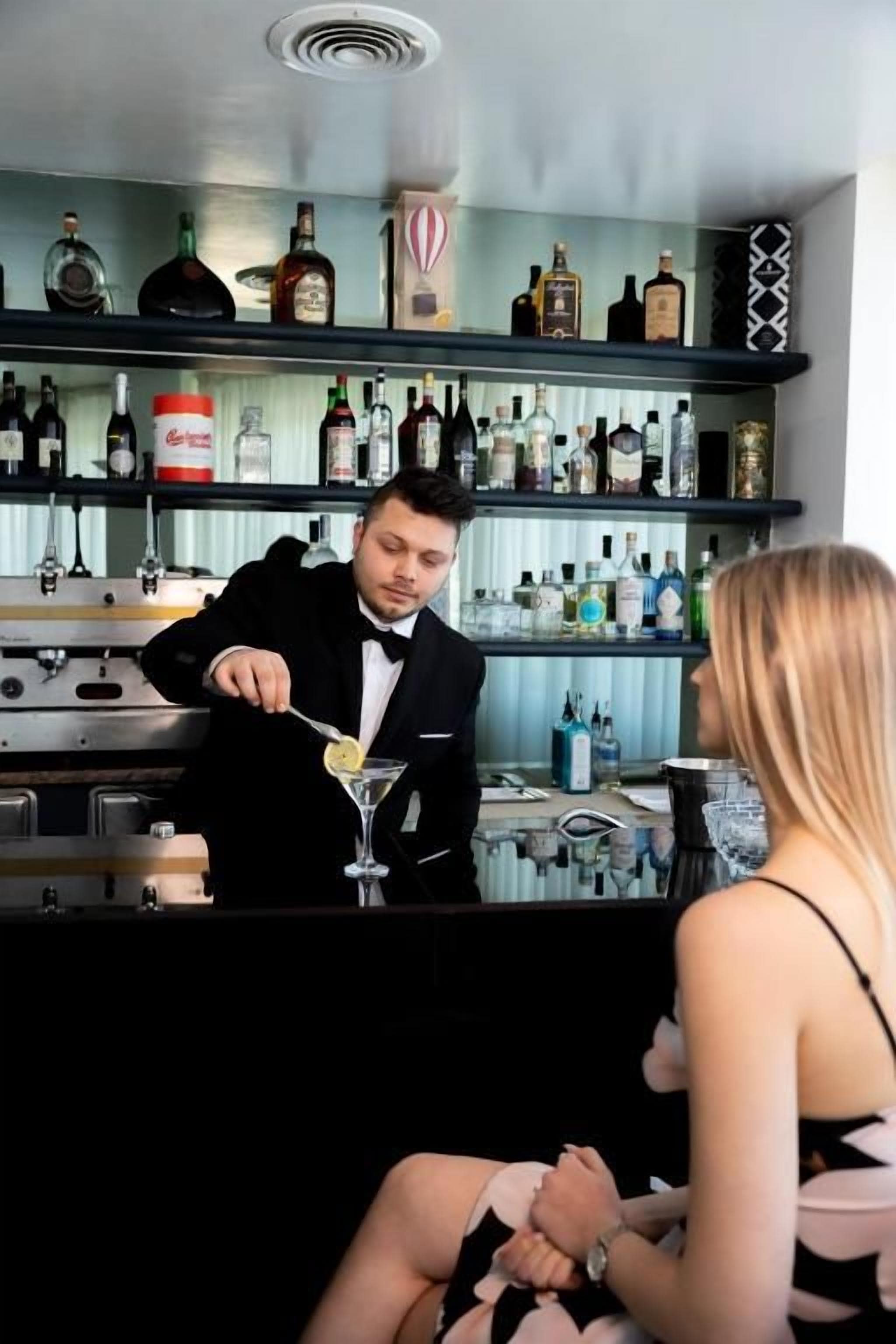 Un bar avec un barman qui prépare un cocktail, des étagères remplies de bouteilles et un comptoir.