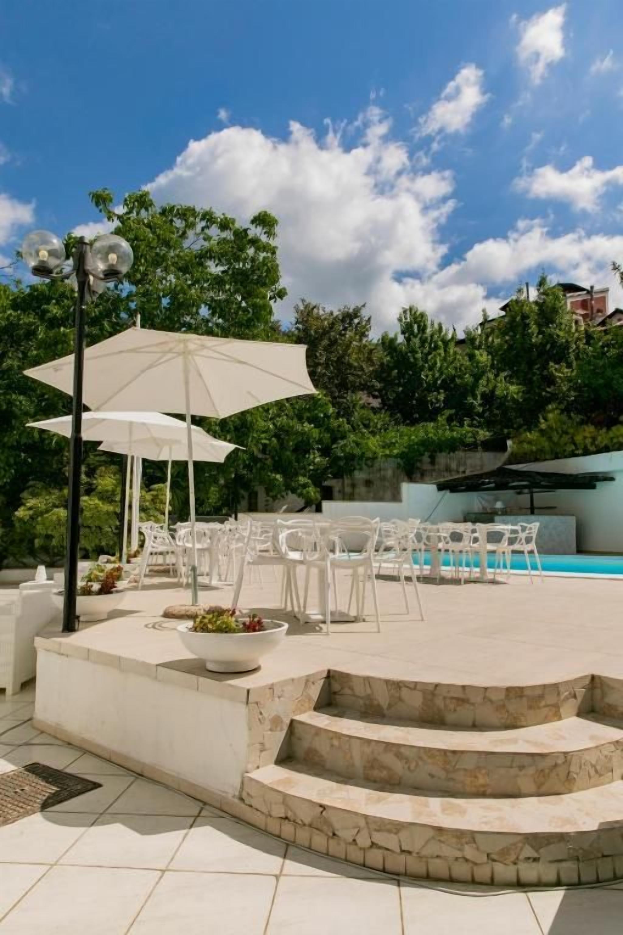 An outdoor seating area with white chairs and a large umbrella, a stone staircase, and a pool in the background.