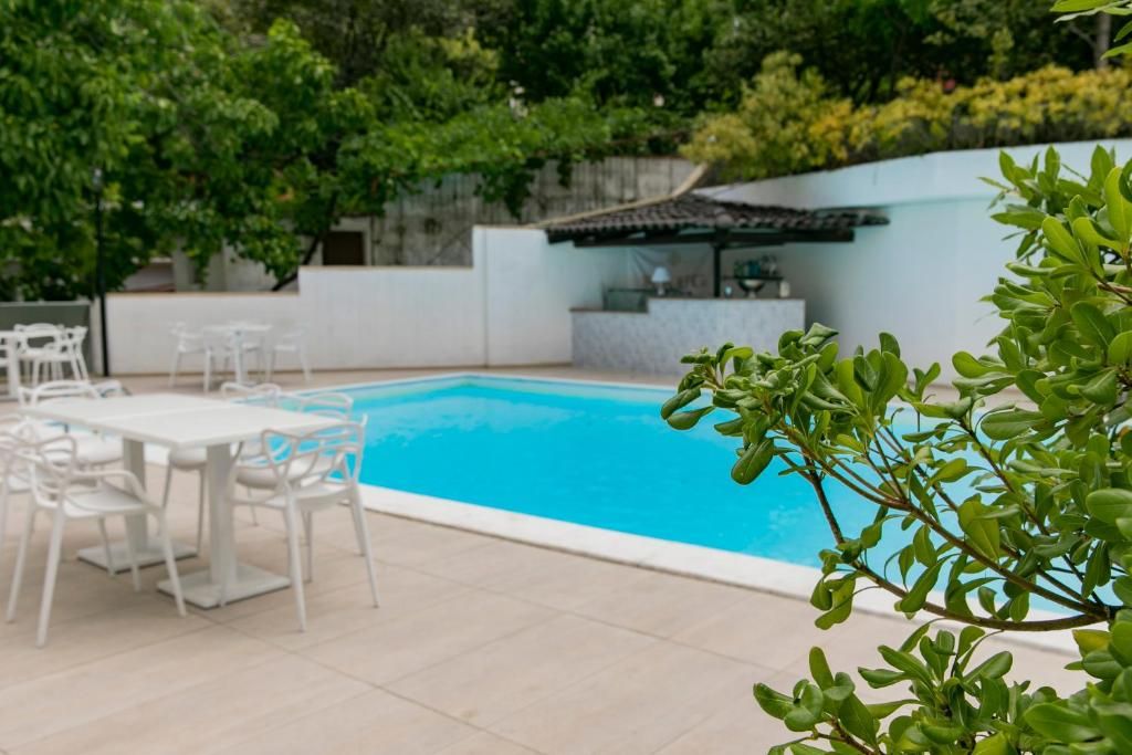 A swimming pool with white chairs and tables, surrounded by greenery and a white wall.