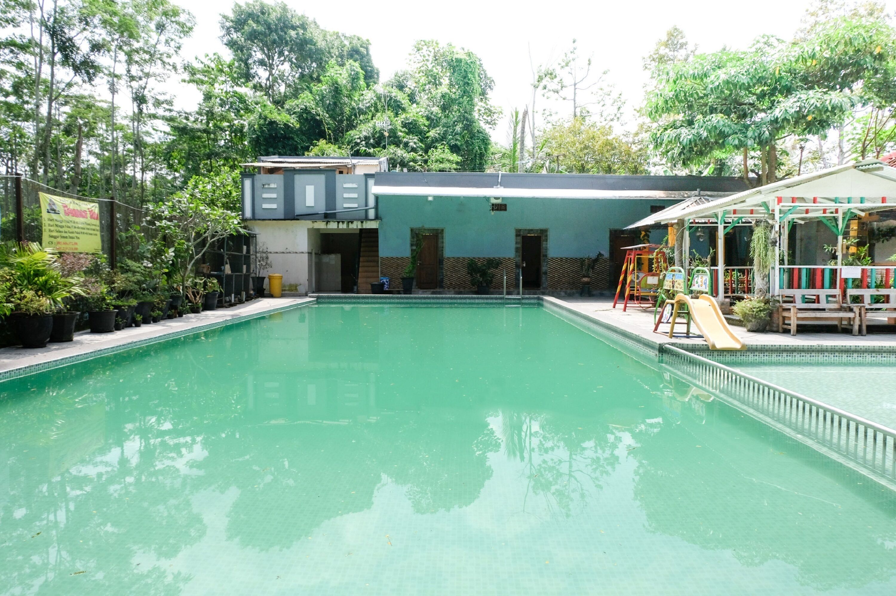 A swimming pool with a slide, surrounded by greenery and a building with a green roof.