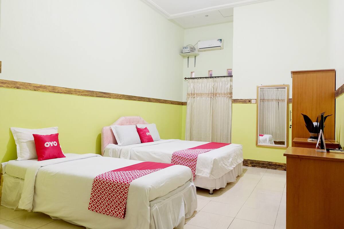 A hotel room with two beds, a wooden desk, a mirror, and an air conditioning unit.