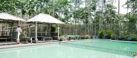 A swimming pool with a tiled deck, surrounded by trees and a covered seating area.