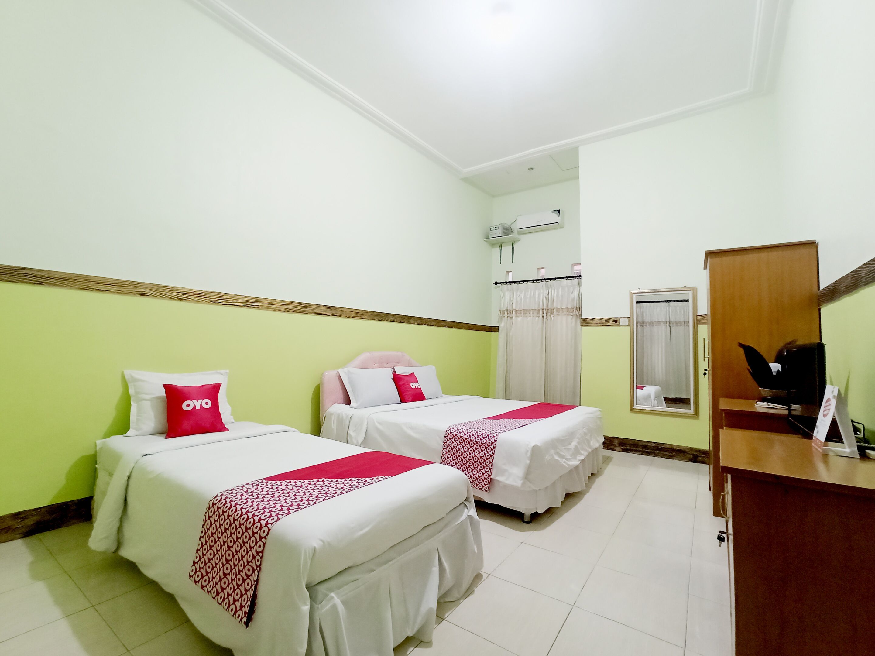 A hotel room with two beds, a wooden desk, a mirror, and an air conditioning unit.