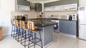 A modern kitchen with grey cabinets, a central island with stools, a built-in washing machine, and a coffee machine. - Flatrock 608 (Cape Town)