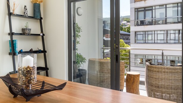 A modern living room with a wooden table, a glass vase with pebbles, a black metal shelf with decorative items, and a view of a balcony with wicker furniture and a building in the background. - Flatrock 608 (Cape Town)