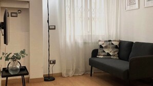 A living room with a dark grey sofa, a small black table with a vase and a plant, a floor lamp, and a window with sheer curtains. - Fiuggi Apartment (Milan)