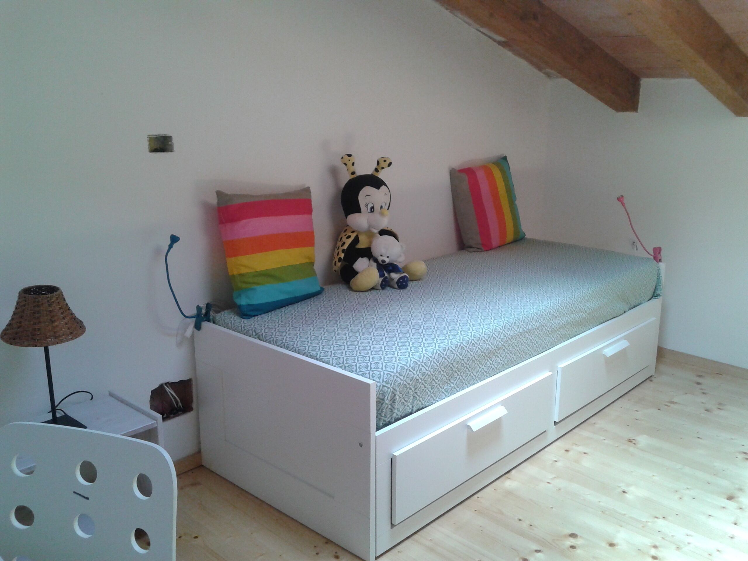 Una habitación pequeña con cama individual, una cama blanca, un cojín a rayas de colores, un peluche y una lámpara.