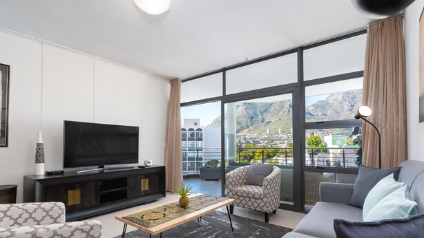 A modern living room with a large TV, comfortable seating, and a view of a cityscape through large windows. - Flatrock 703 (Cape Town)