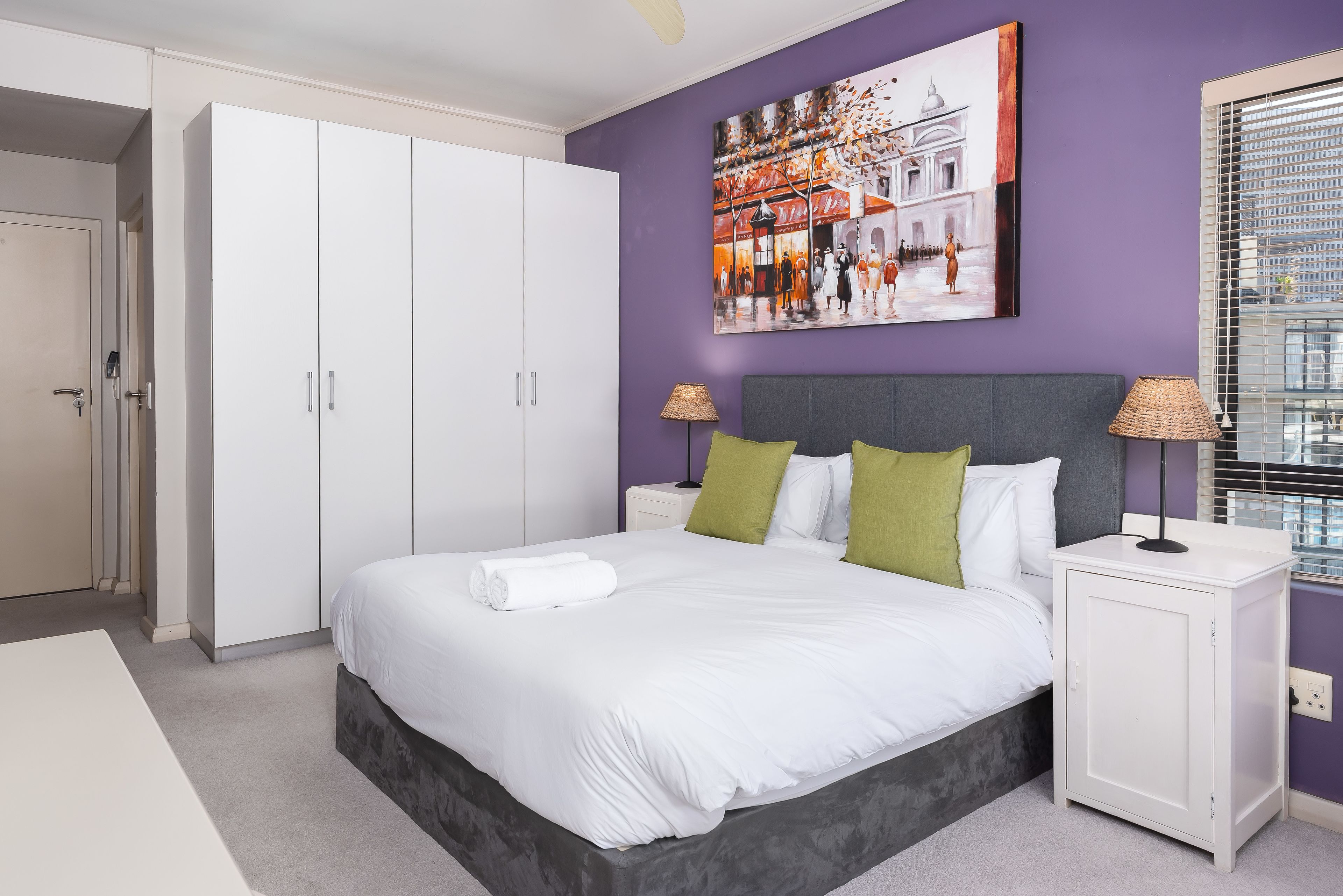A modern bedroom with a large bed, white wardrobe, and a cityscape painting on the wall.
