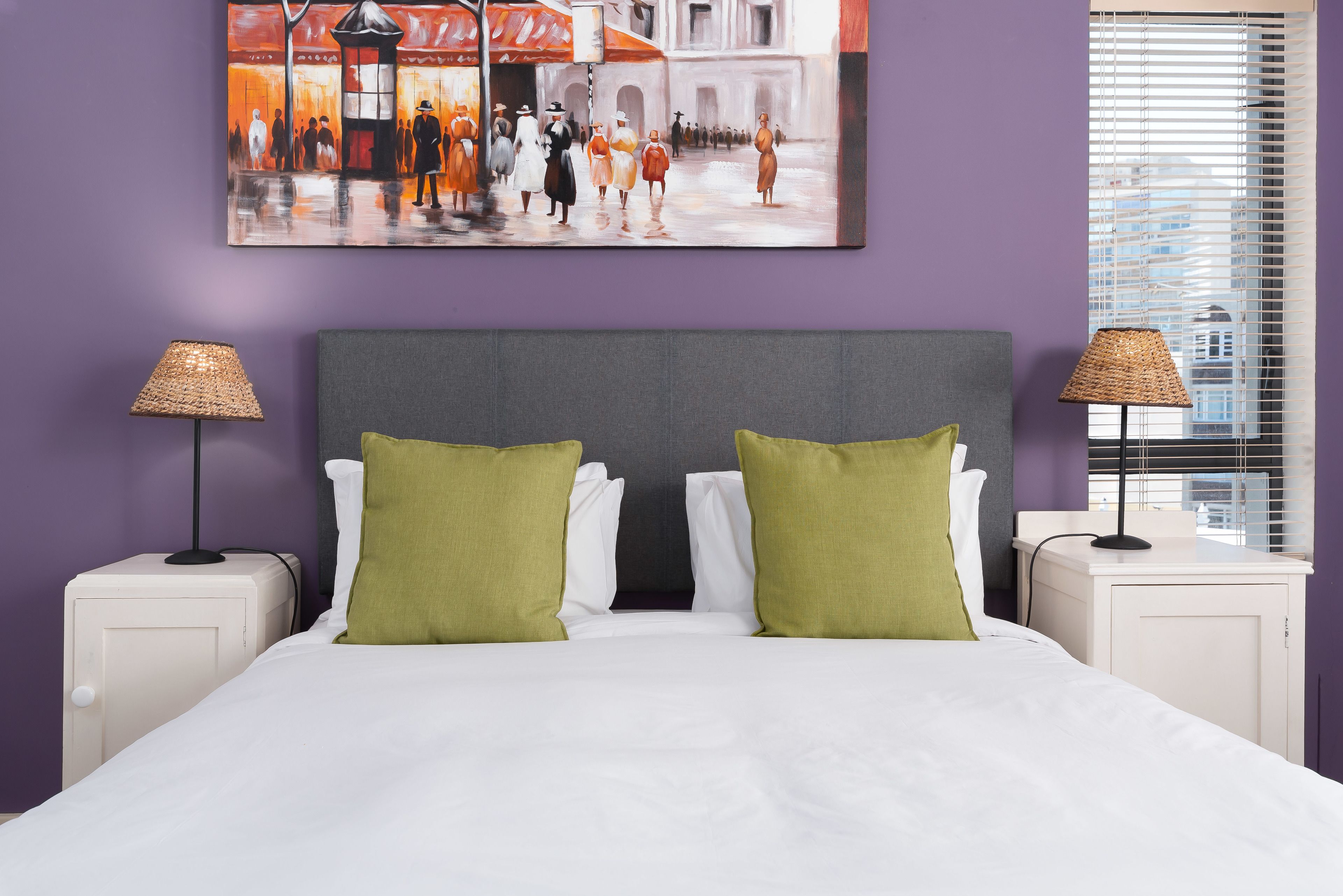 Een netjes opgemaakt bed met groene kussens, een schilderij erboven en twee bedlampjes.