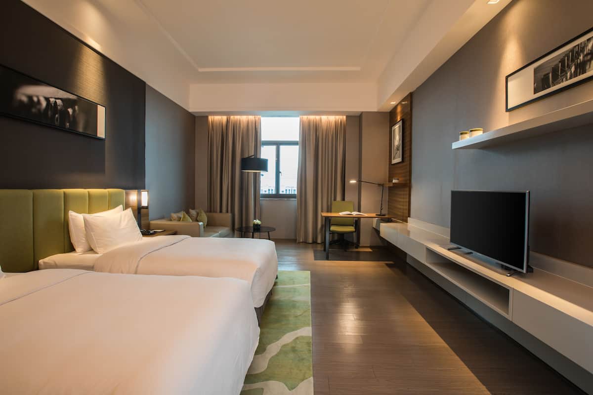 A hotel room with two beds, a TV, a desk, and a window with curtains.