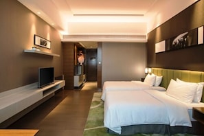 Ein Hotelzimmer mit zwei Betten, einem Flachbildfernseher, einem Regal mit gerahmten Fotos und einem Gang, der in ein weiteres Zimmer führt.