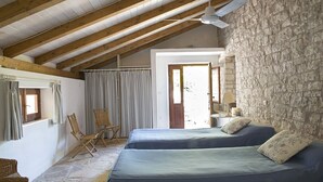 A bedroom with two beds, wooden beams, stone walls, a ceiling fan, and a window with curtains.