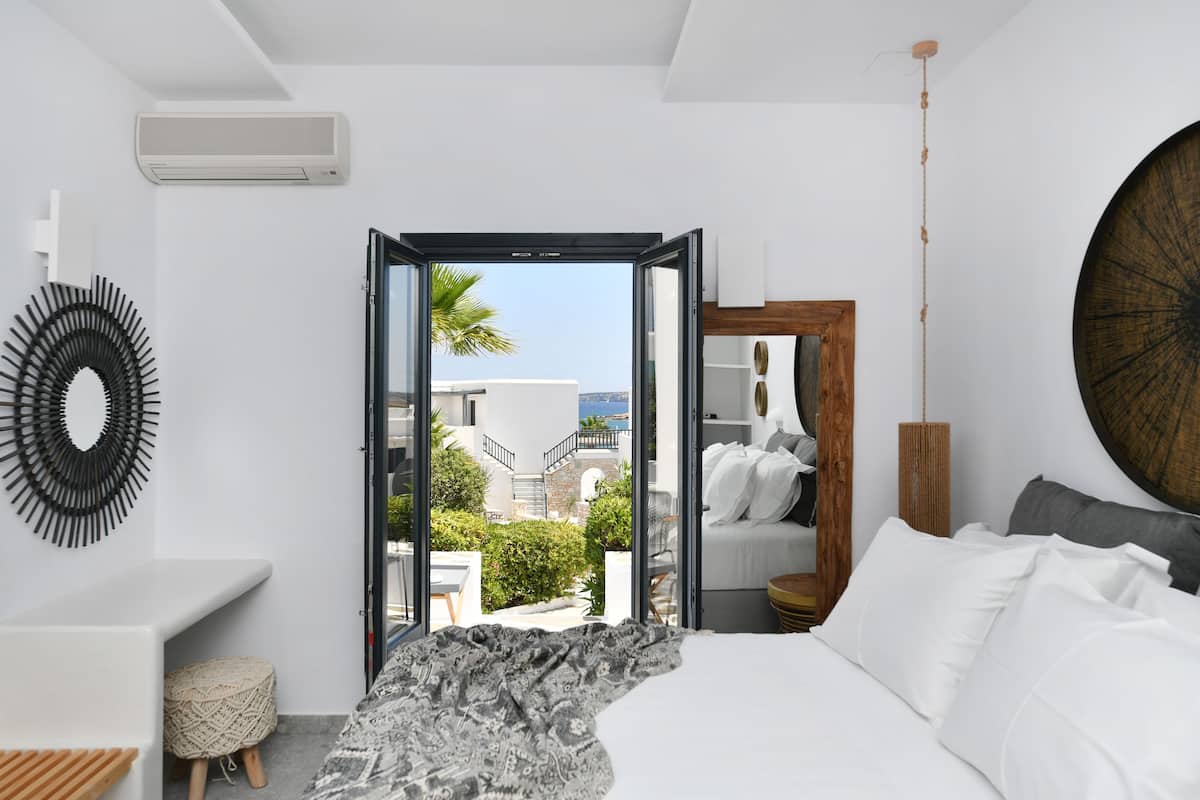 A modern bedroom with a bed, a bedside table, a mirror, and a view of a balcony with a palm tree.