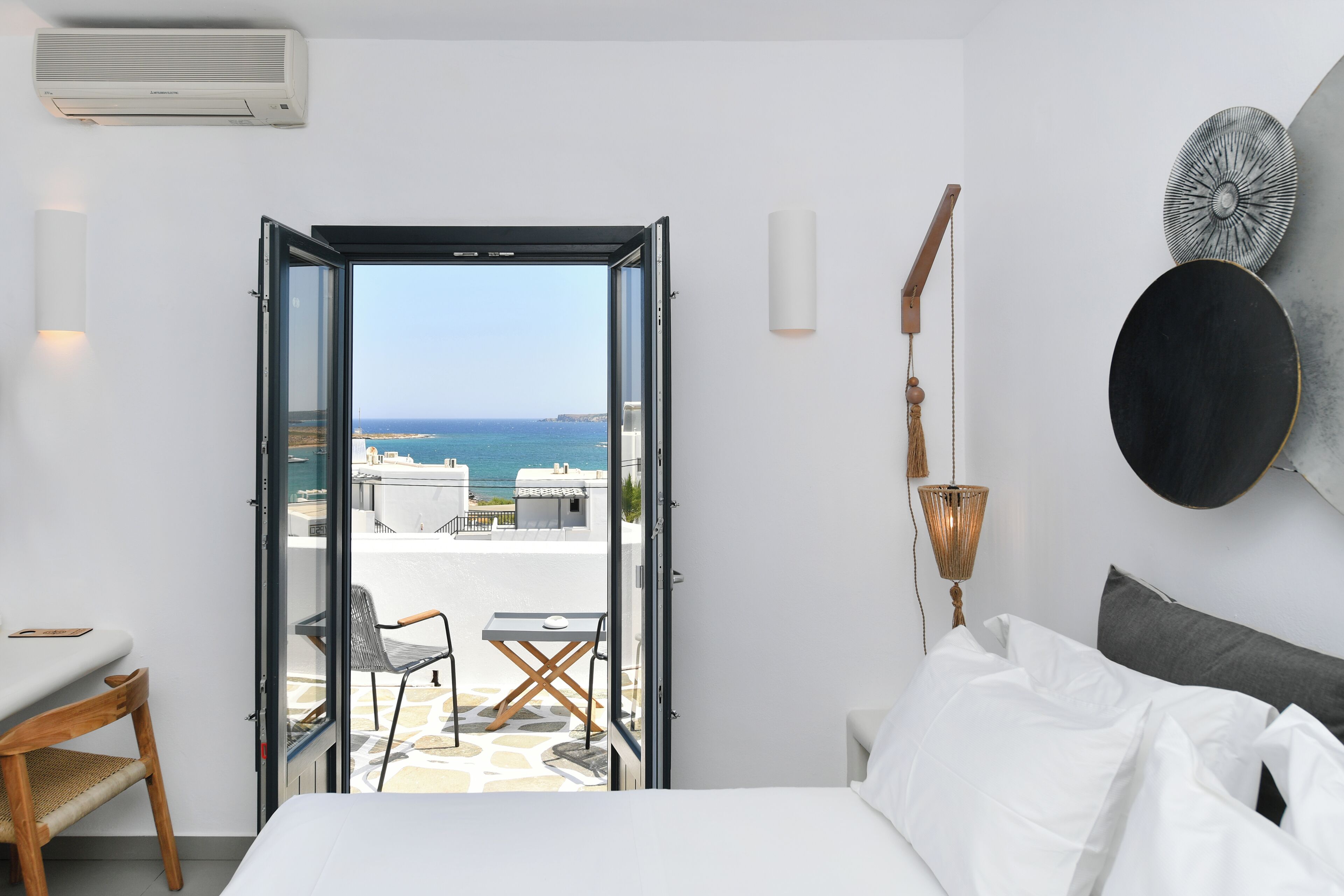 A hotel room with a bed, a desk, and a chair. There is a balcony with a view of the sea.