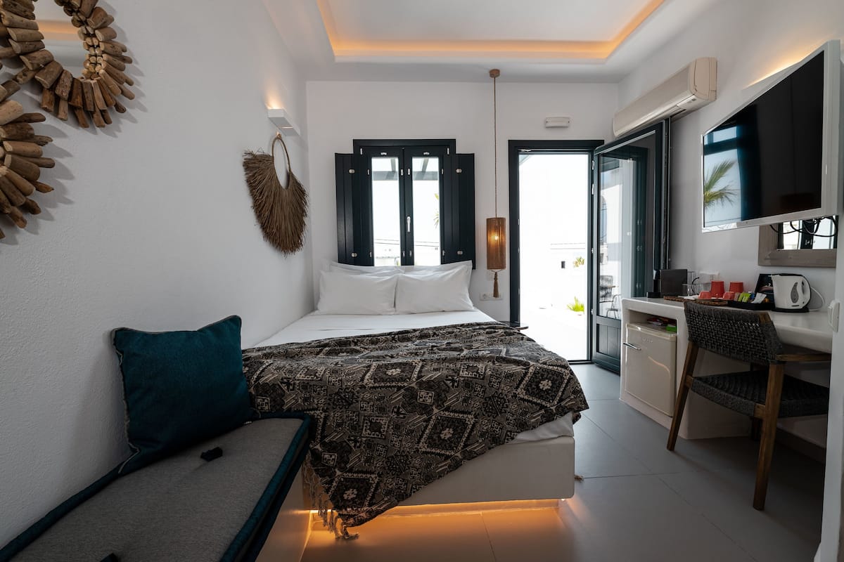 A modern bedroom with a bed, a desk, and a chair. There is a window with shutters, a wall-mounted TV, and a door leading to another room.