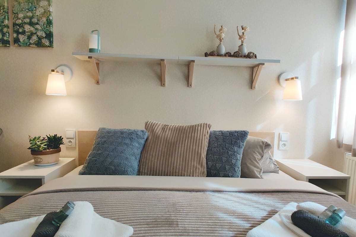 A hotel room with a large bed, two bedside tables, a nightstand, a wall-mounted shelf with decorative items, and a potted plant.